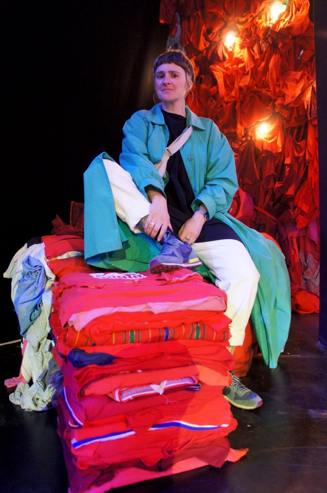 Photo of Sexy Roy sitting on piles of fabric. They are wearing a long turquoise coat and wearing big hooped ear rings. Behind them is a wall of illuminated red fabric.