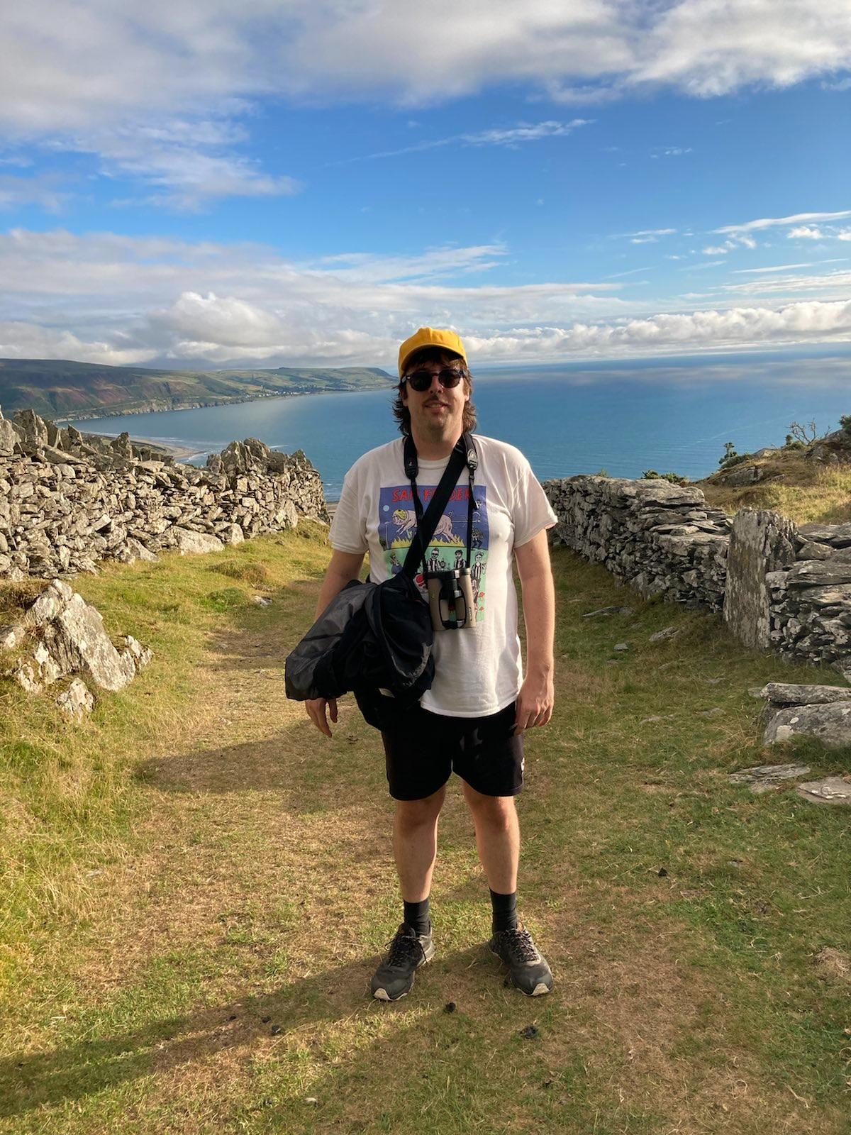 Photo of DJ Superstar DJ out in the Welsh mountains. They are wearing a Sam Fender t-shirt and have binoculars around their neck. It's sunny and there's beautiful sea and mountain in the background.